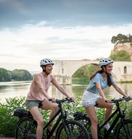 Two people wearing helmets ride bicycles along a scenic riverside path. A historic stone bridge arches over the river in the background, under a cloudy sky.