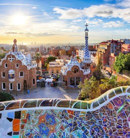 Sunlit view of Park Güell in Barcelona, showcasing colorful mosaic benches, whimsical architecture, and a vibrant skyline under a bright blue sky.